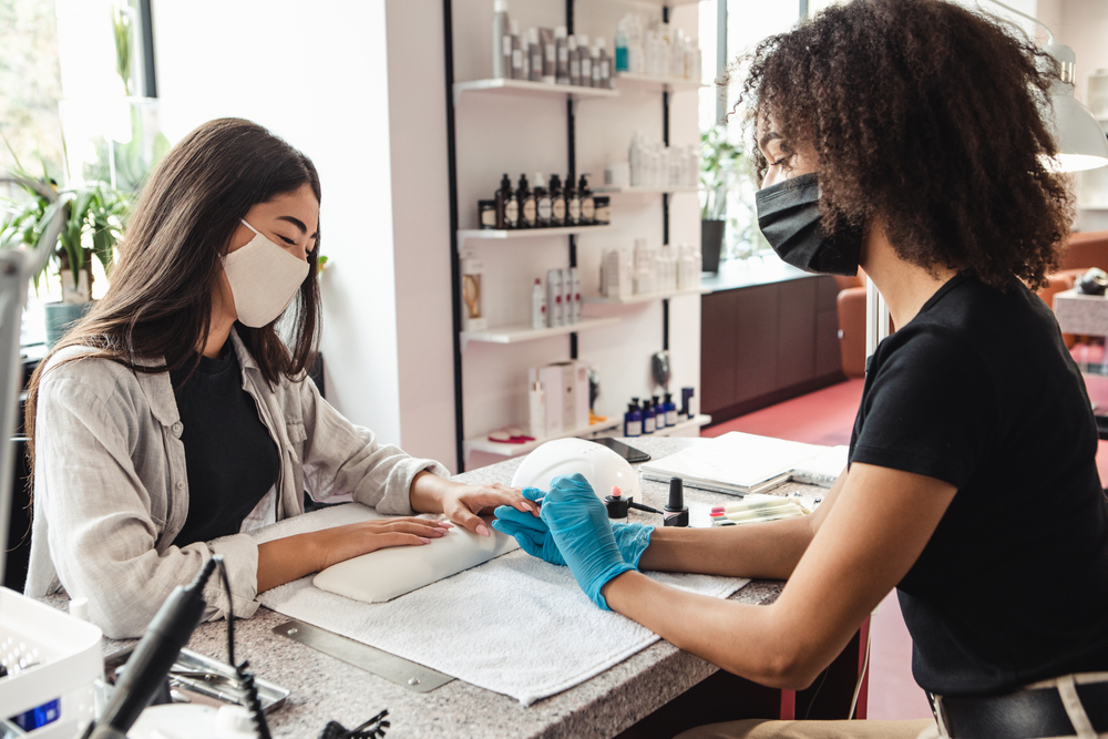 Returning to work after quarantine and gossip at nail salon. African american woman in protective mask files nails with electric device to smiling asian lady client in studio interior