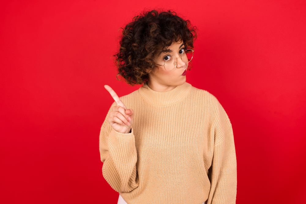 No sign gesture. Closeup portrait unhappy Young beautiful Arab woman wearing knitted sweater standing against red background raising fore finger up saying no. Negative emotions facial expressions.
