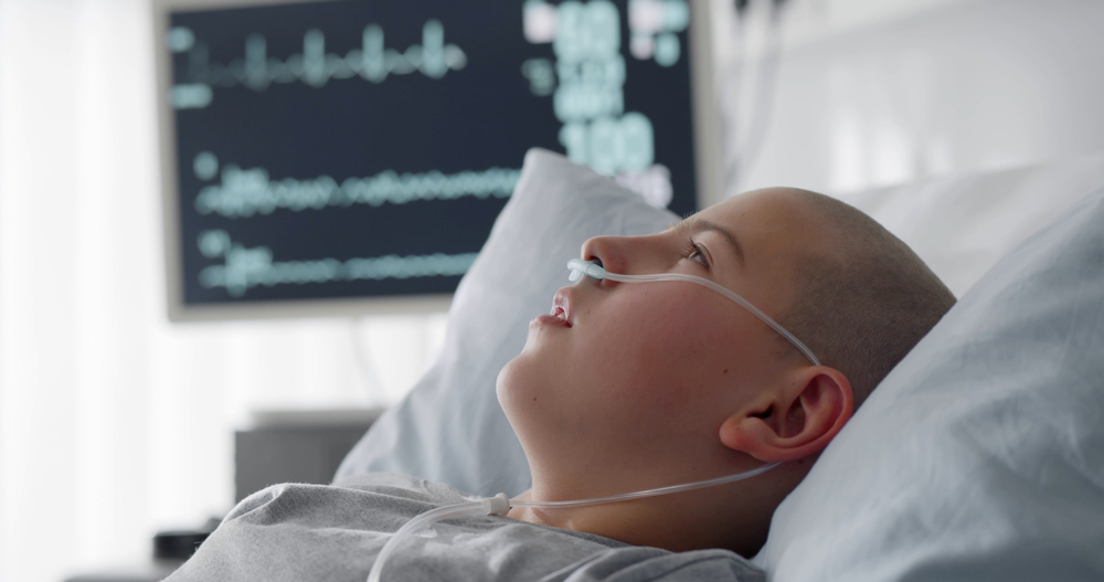Sick child with cancer lying in hospital bed with nasal cannula. Close up portrait of bald teen boy patient resting in bed after chemotherapy treatment or surgery