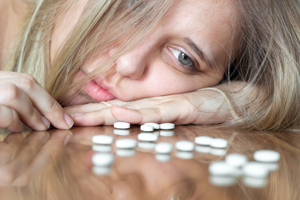 A young caucasian beautiful upset blonde woman puts her head on the table and looks at the antidepressants. Despair, apathy, depression, frustration, exhaustion, thoughts of suicide, mental disorder
