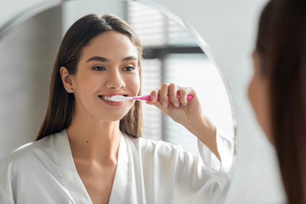 Morning Hygiene. Portrait Of Attractive Young Lady Brushing Teeth Near Mirror, Happy Beautiful Woman Smiling To Her Reflection While Standing In Bathroom With Toothbrush In Hand, Selective Focus
