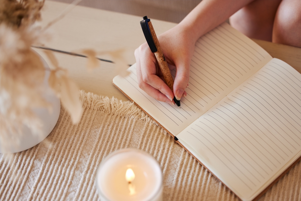 Hand, candle and woman writing in journal with top view for calm, peace mindset and relax morning routine in home. Hands, notebook and diary planning goals, idea vision or creative writer lifestyle
