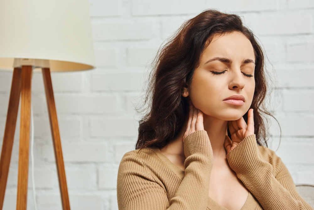 Young brunette woman closing eyes during self-massage of lymphatic system and thyroid gland function support at home, self-care ritual and holistic wellness practices concept, tension relief