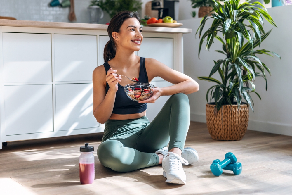Shot of athletic woman eating a healthy bowl of muesli with fruit sitting on floor in the kitchen at home
