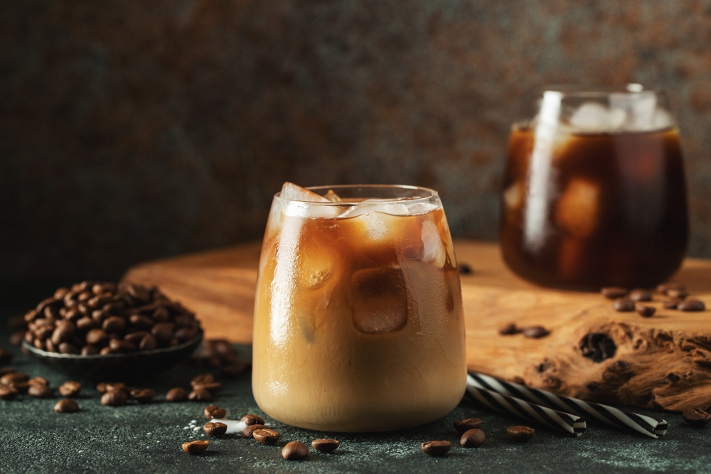 Ice coffee in a tall glass with cream poured over, ice cubes and beans on a dark concrete table. Cold summer drink with tubes on a black background with copy space.