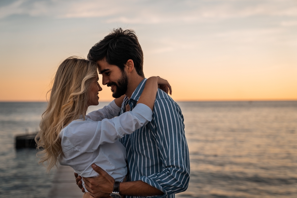Lovely couple bonding at the beach in summer. Romantic male and female looking each other into eyes during the sunset. Beautiful couple in love standing face to face.