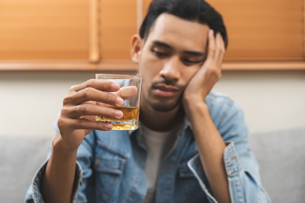 Health care alcoholism drunk, fatigue asian young man hand holding glass of whiskey, alone depressed male drink booze on sofa at home. Treatment of alcohol addiction, suffer abuse problem alcoholism.
