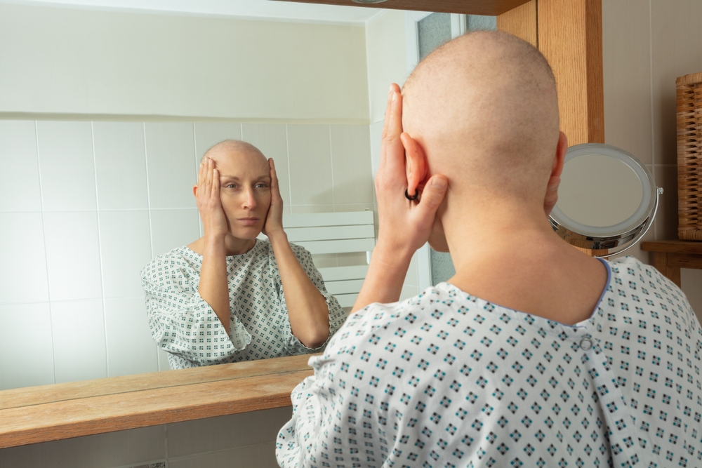 A bald woman in a hospital gown examines her reflection in a bathroom mirror