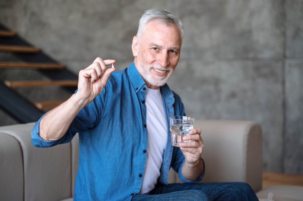 Happy smiling senior man taking supplements, daily vitamins for healthy lifestyle. Male holding tablet and glass with water. Intaking capsule with omega3, vitamin D