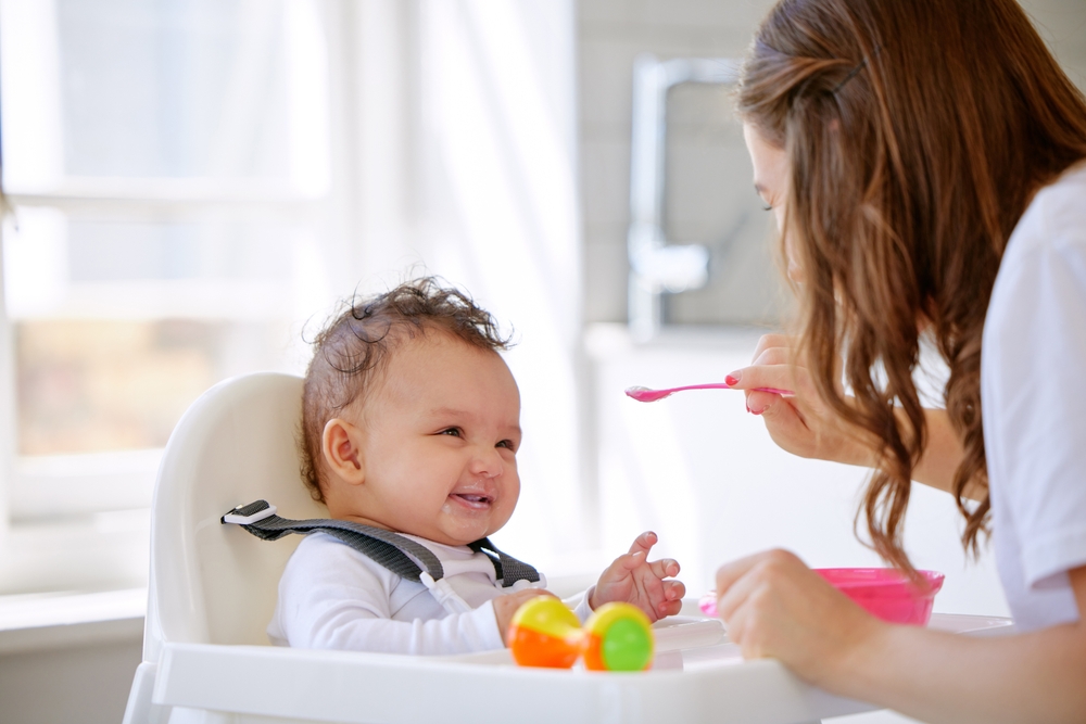 Mother, kitchen and feeding baby with high chair for meal, snack time or breakfast at home. Happy mom with infant, newborn or child for food, nutrition or care with love, eating or support at house