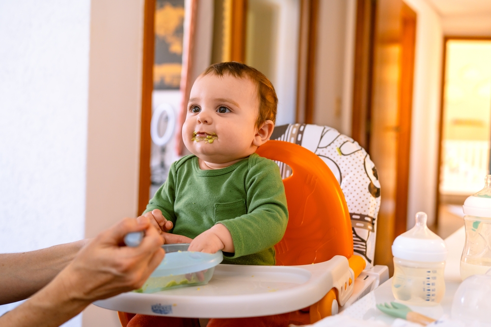 Smiling baby boy in green outfit being spoon-fed avocado. Adorable toddler enjoying a healthy meal with a playful expression at home.