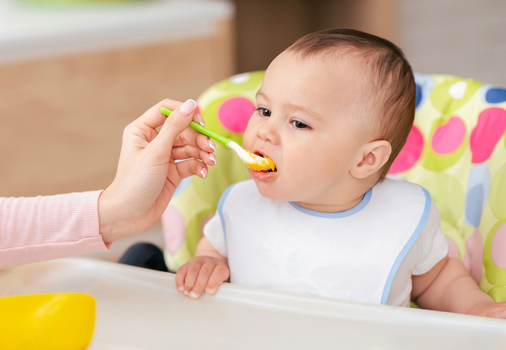 A mother is feeding her baby boy at the kitchen table, with him seated in a high chair. The mood is nurturing and loving, with a homely kitchen setting.