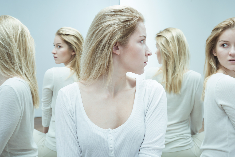 Shot of a young woman dressed in white, looking back with anxiety at her reflections in the mirror
