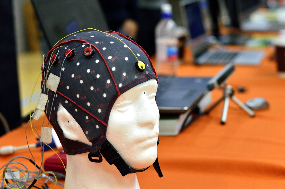 The electroencephalogram (EEG) head cap with flat metal discs (electrodes) attached to a white plastic model’s head shown in a science exhibition, with laptops blurred at the background.
