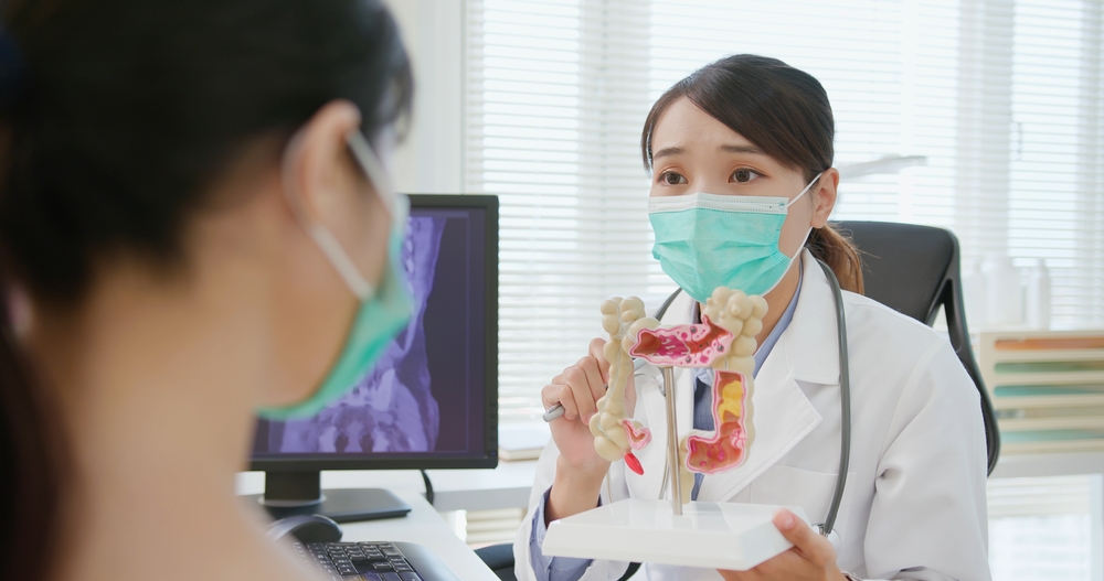 asian woman wears face mask to prevent COVID19 has colorectal cancer diagnosis in hospital - female doctor showing colonoscopy results and xray to patient on computer and explain by anatomical model
