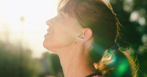 young woman smiling looking up at sky, sun shining through trees in background