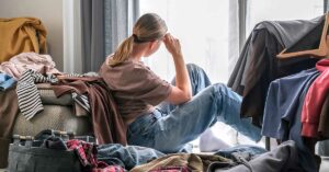 30 Essential Things to Declutter for a Healthier Mind and Body woman sitting in a room full of clutter. Declutter concept