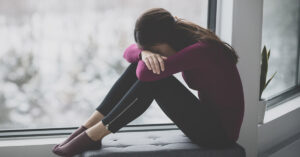 12 Warning Signs of Narcissistic Victim Syndrome depressed woman sitting by a window with head resting over arms on knees