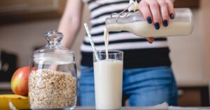 22 Calcium-Rich Alternatives to Milk That Are Just as Beneficial woman wearing blue jeans and a striped shirt pouring glass of milk from glass bottle into a glass with a straw in it