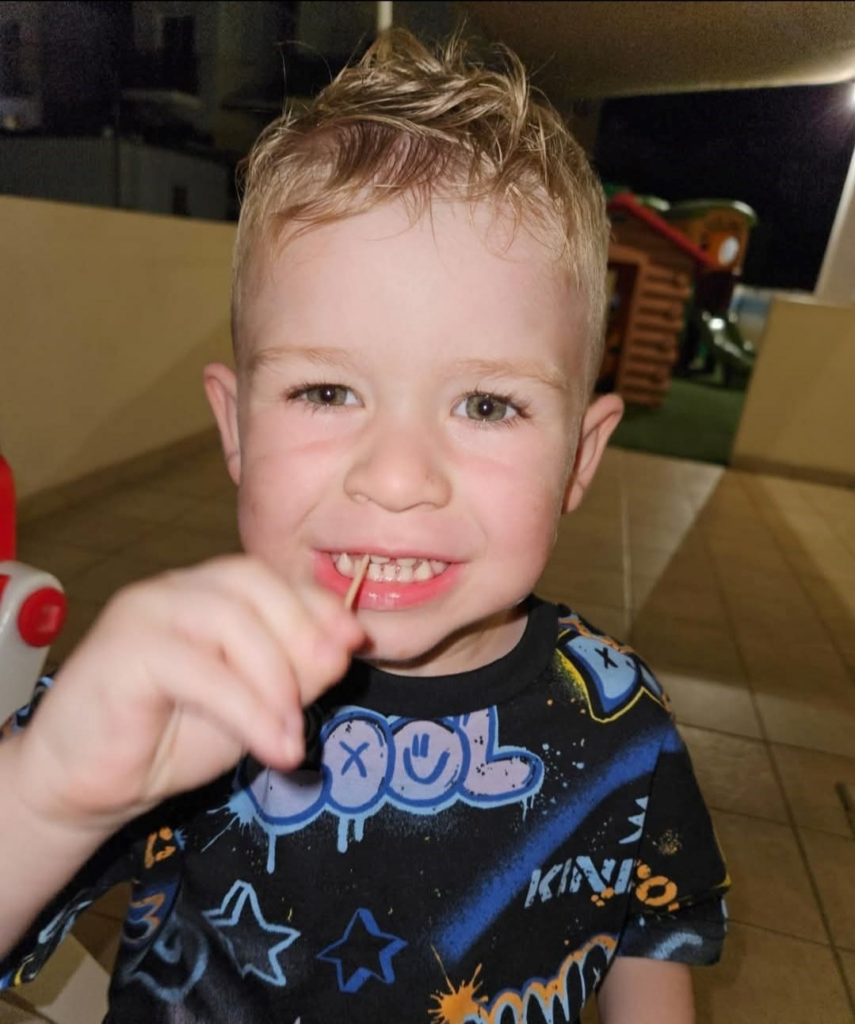 A heartwarming photo of Jaxon, a young boy with blonde hair and a bright smile, holding a toothbrush. This image captures the innocence and joy of childhood, serving as a poignant reminder of how meningitis symptoms can rapidly progress.