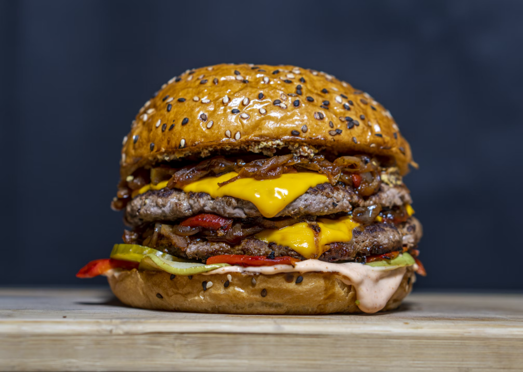 Gourmet burger with multiple layers, melted cheese and sauce on a seeded bun - representing the complex engineered foods that food scientist Dr. Barry Smith once helped develop before discovering the health impacts of ultra-processed foods.