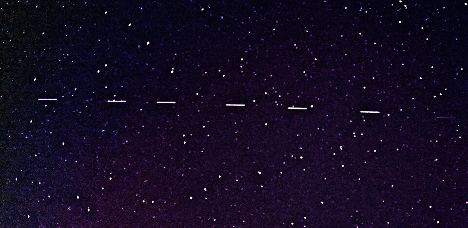A row of Starlink satellites moves across a star-filled night sky. The satellites appear as short, bright lines in a straight path. This is part of SpaceX's plan to provide internet using satellites in low Earth orbit.