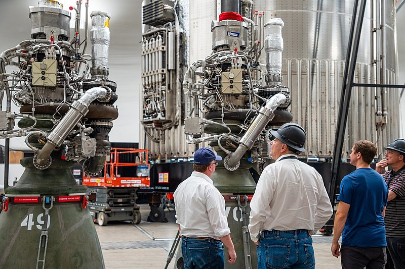 SpaceX engineers stand near large Raptor rocket engines. They are checking the setup inside a testing facility. These engines are used in Starship flights.