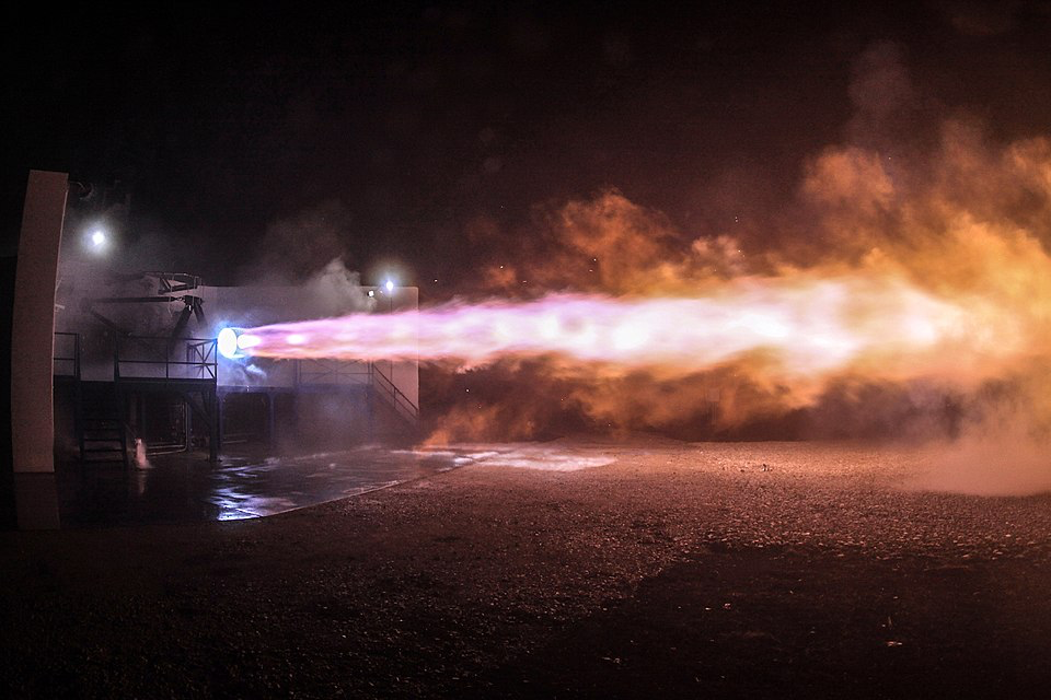 A Raptor engine test fires at night. Bright flames shoot out as part of the engine testing process. This engine powers the upper stage of Starship.