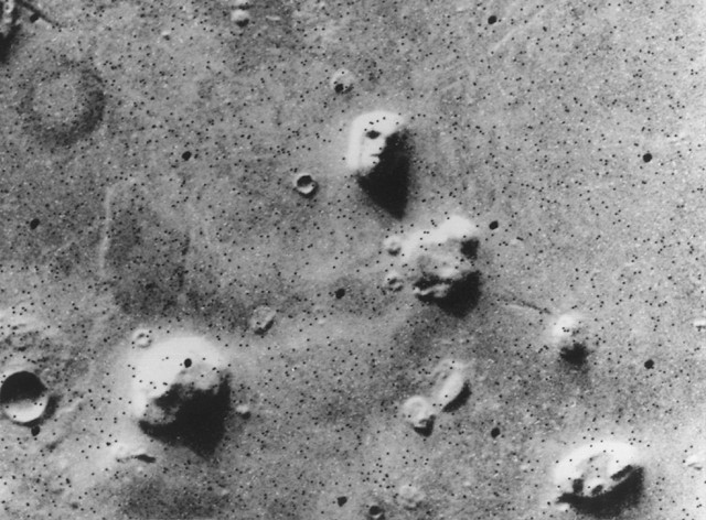 Viking orbiter photograph from the 1970s showing the controversial "Face on Mars" formation in the Cydonia region, along with surrounding pyramid-like structures. The image quality is grainy with high contrast, making the formations appear to have distinct geometric shapes that sparked theories about ancient Martian civilization ruins.