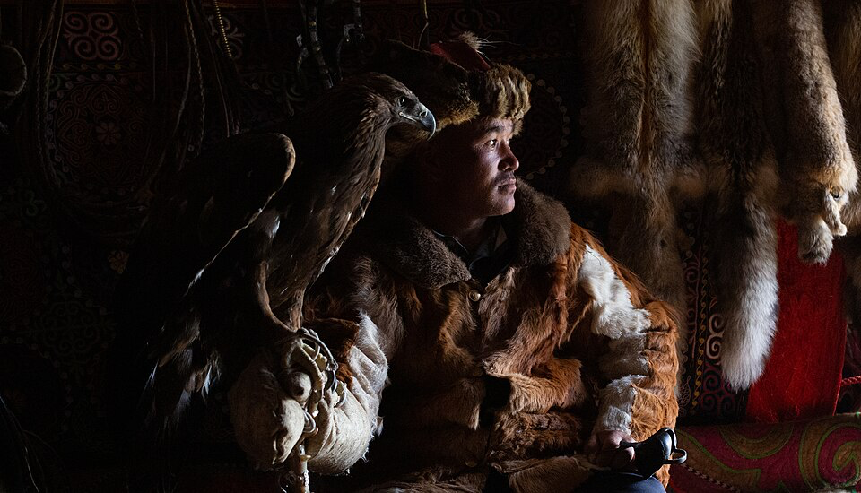 Traditional Kyrgyz eagle hunter in traditional fur clothing holding a trained bird of prey inside a yurt, with animal pelts visible in the background.