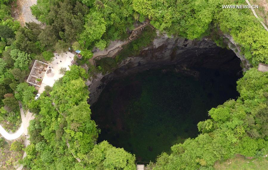 Aerial view of a massive karst sinkhole in Guangxi, China, revealing a circular abyss surrounded by lush green forest with small structures visible at the rim for scale.