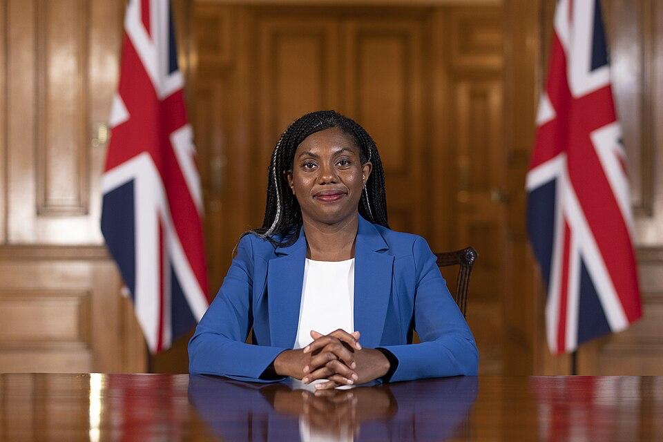 Conservative leader Kemi Badenoch sits at a wooden table in a government meeting room, wearing a blue blazer over a white top. She has her hands clasped in front of her with two UK flags visible in the background.