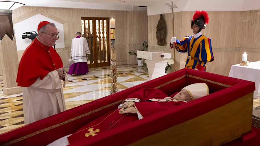 Pope Francis's body lies in state in a simple wooden casket with red interior lining, dressed in traditional red papal vestments. A rosary rests on his folded hands, and a small crucifix is placed on his chest. A cardinal in red robes stands solemnly beside the casket, while a Swiss Guard in traditional colorful uniform stands watch. The scene takes place inside a marble-lined room within the Vatican.