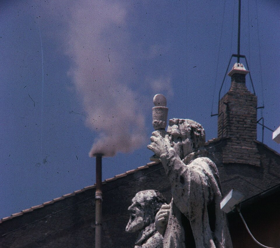 White smoke rises from a chimney atop the Vatican, signaling the election of a new pope during the 1963 conclave that selected Pope Paul VI. The image shows the historic smoke against a blue sky, with a stone statue of a saint or apostle visible in the foreground. This traditional signal announces to the world that the College of Cardinals has successfully chosen a successor to lead the Catholic Church.