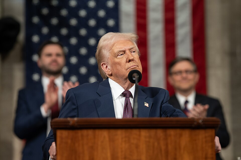 President Donald Trump at a podium with the American flag in the background, whose tariff policies have been challenged by Governor Newsom