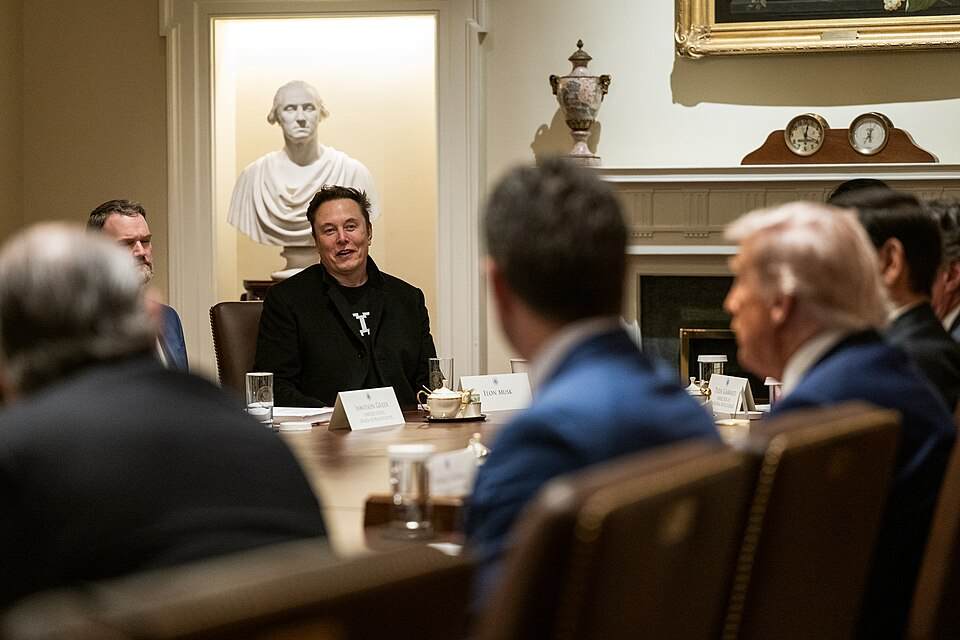 A cabinet meeting at the White House featuring Elon Musk seated at the table wearing dark clothing with a white statue visible in the background, alongside traditional White House decor.