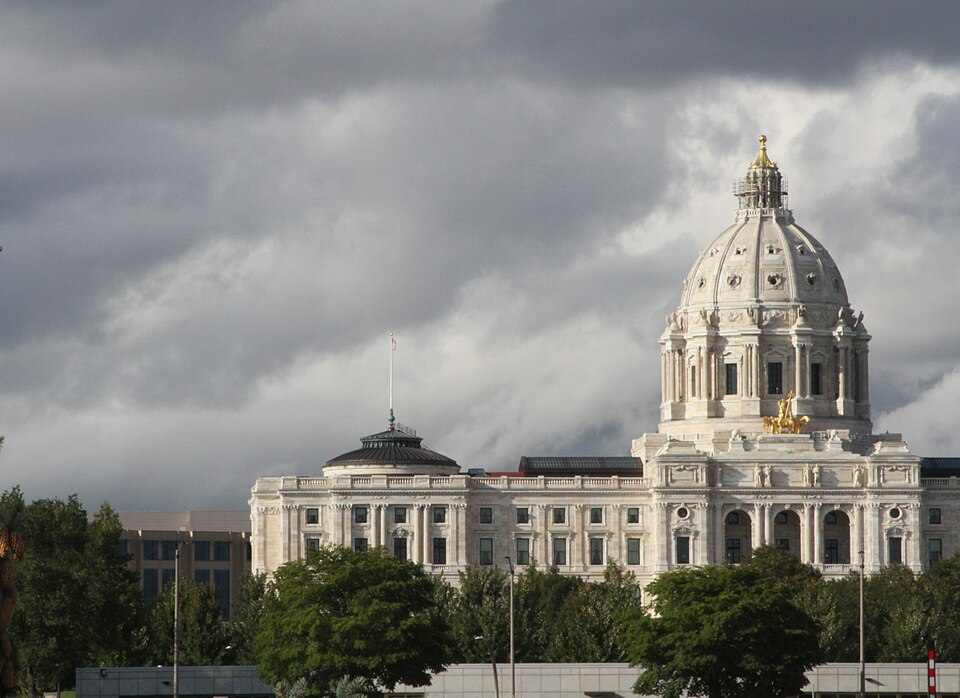 The Minnesota State Capitol building with its distinctive white dome and golden details standing prominently against dramatic storm clouds, creating a contrast between the bright architecture and the moody sky.