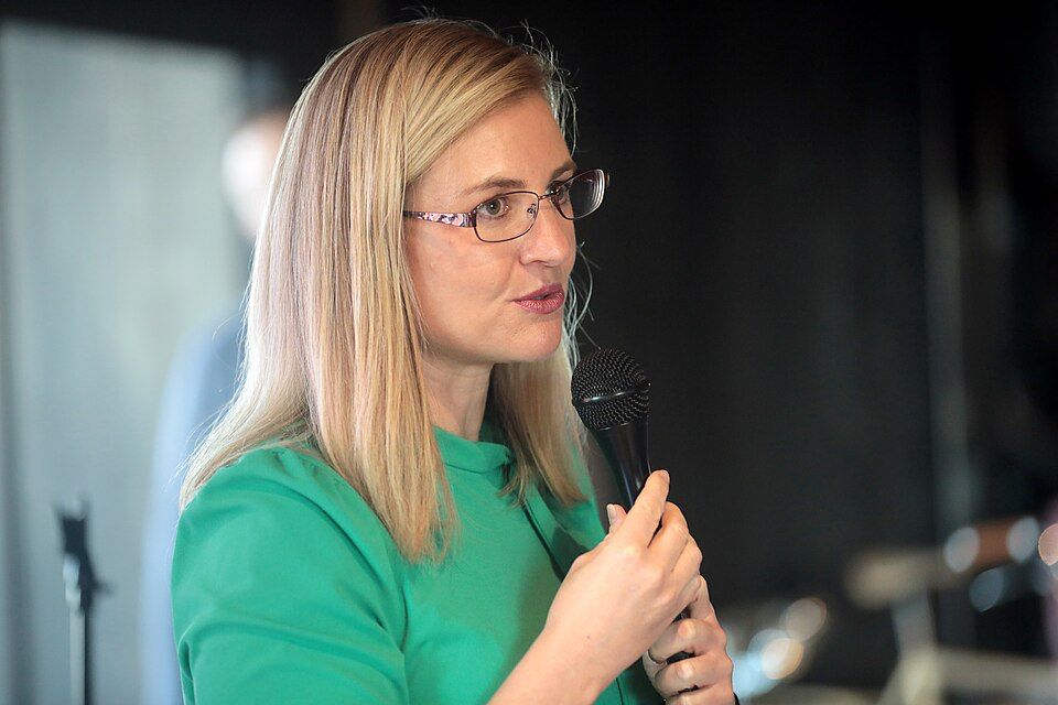 Phoenix Mayor Kate Gallego wearing glasses and a green top speaking into a microphone, advocating for residents to obtain their Real ID before the deadline.