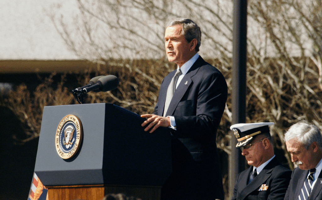 President George W. Bush speaking at an official podium with the presidential seal, who signed the Real ID Act into law in 2005 following 9/11 Commission recommendations.