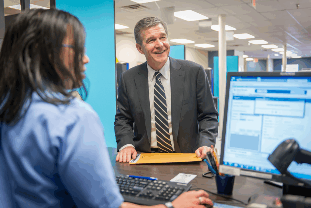 Customer service interaction at a counter with a computer terminal, showing a person in a blue shirt speaking with a representative about identification requirements.
