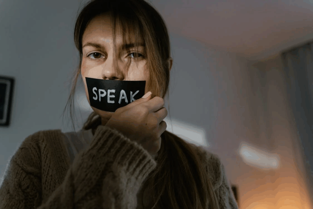 A woman with long hair holding a black tape with the word 'SPEAK' over her mouth, symbolizing silenced victims finding their voice.