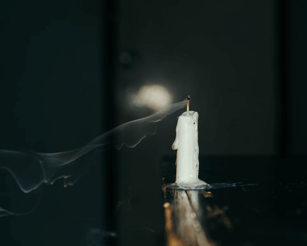 A white candle burning in darkness with wispy smoke, representing remembrance and the search for truth.