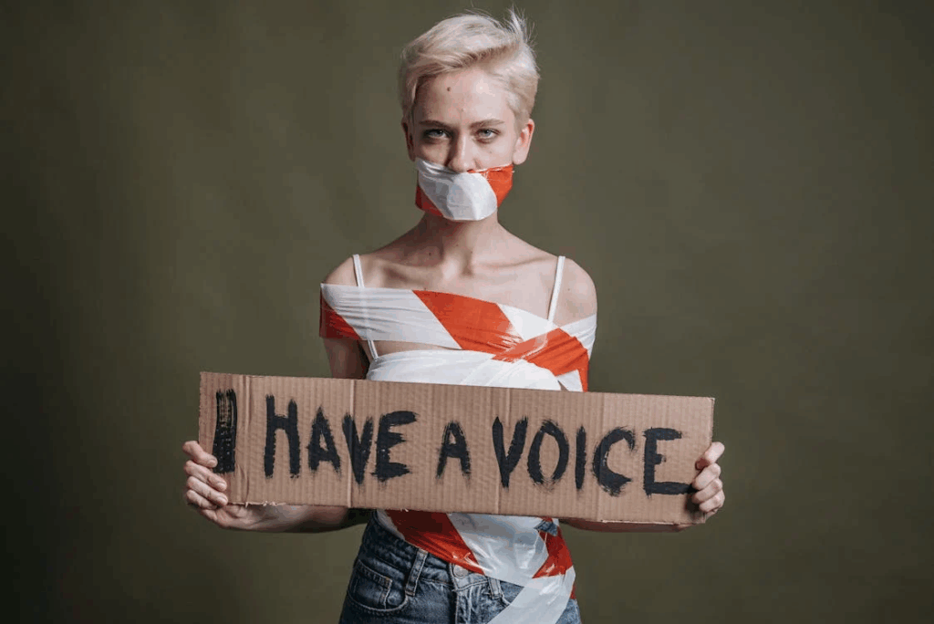 A person with short blonde hair wearing red and white tape across their mouth while holding a cardboard sign reading 'I HAVE A VOICE', representing Virginia Giuffre's fight for justice.
