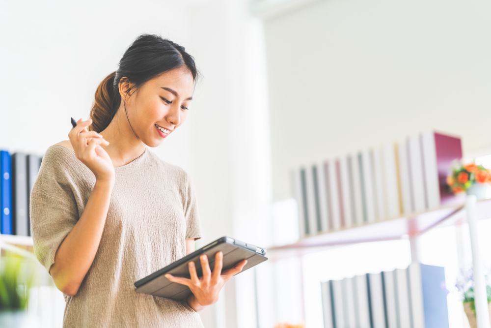 Young beautiful Asian girl working at home office using digital tablet, with copy space. Business owner entrepreneur, small business startup company, or casual freelance job lifestyle concept.