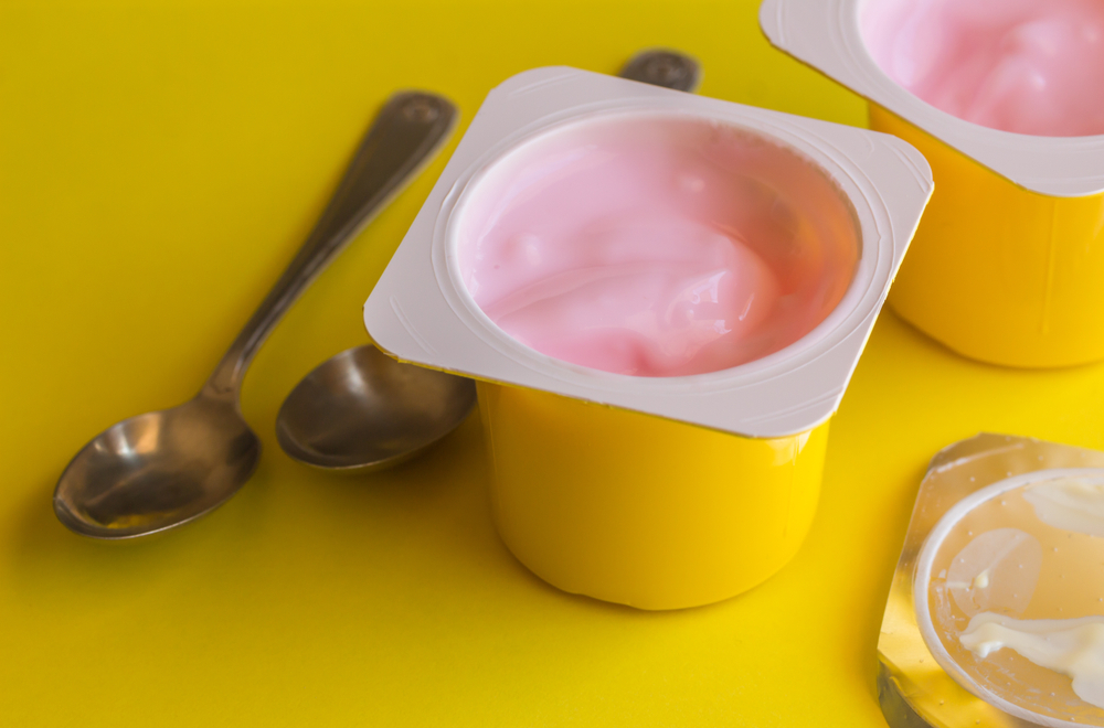 Two strawberry yogurt cups with small silver spoons on bright yellow background - pink yoghurt pots