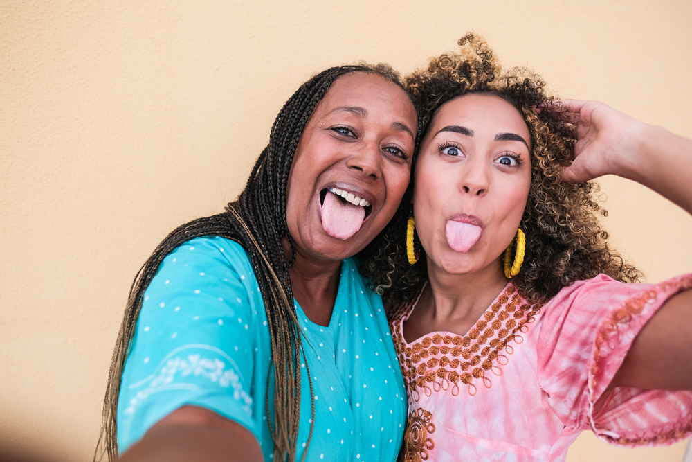 Beautiful african mother and adult daughter stick their tongue out while they take a selfie - Family love