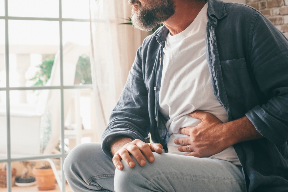 Man suffering with severe stomach pain sitting at home. Hand of mature guy holding abdomen suffering from ache, diarrhea or indigestive problem. Caucasian guy pressing on belly on painful sensation