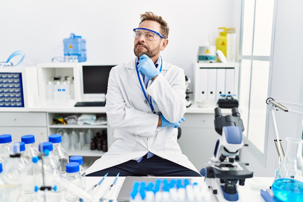 Middle age man working at scientist laboratory serious face thinking about question with hand on chin, thoughtful about confusing idea