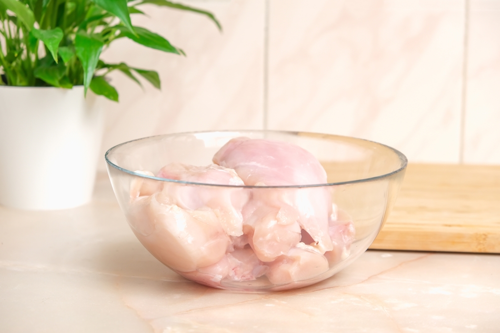 Defrosted raw chicken fillet in a glass bowl. Fresh raw chicken breast for cooking chopped chicken cutlets. Glass bowl and wooden cutting board for slicing chicken fillet.