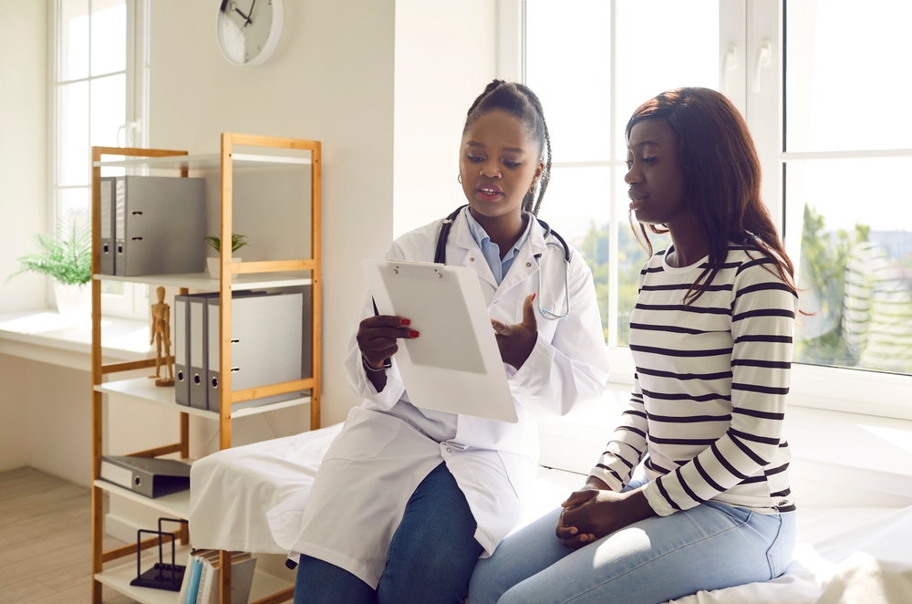 Young doctor woman holding report file with appointment and prescribe treatment to a female african american sick worried patient sitting on the couch in clinic during medical examination.